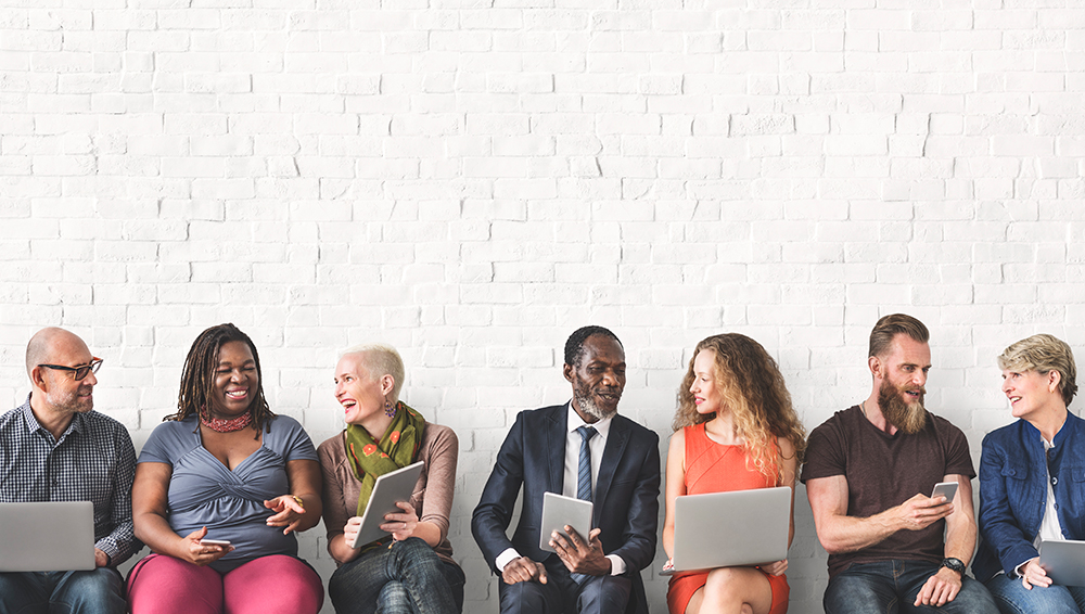 Blog Default Image. Group of people sitting against a wall sharing things on electronic devices.
