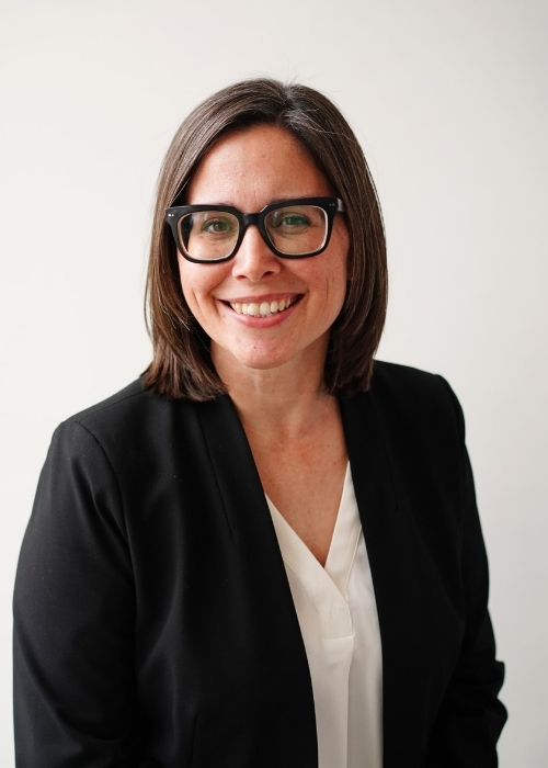 a smiling woman posed against a white wall wearing a white blouse and a black blazer