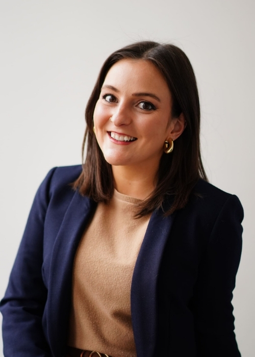 a smiling woman posed against a white wall wearing a tan sweater and a navy blazer